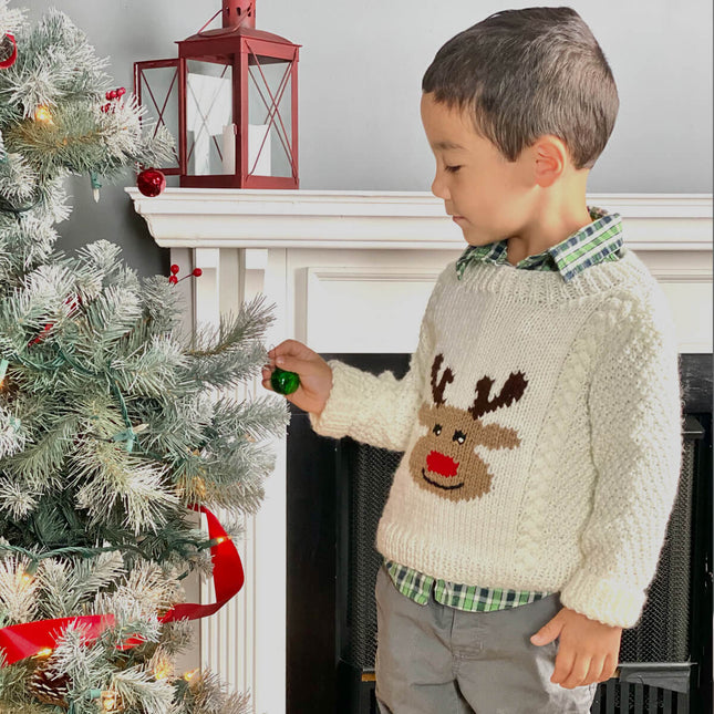 Child hanging ornaments on christmas tree wearing a knit reindeer sweater with aran cables