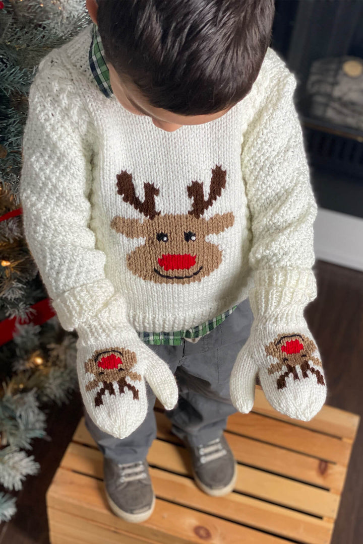 Child hanging ornaments on christmas tree wearing a knit reindeer sweater with aran cables and knit reindeer mittens